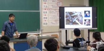 教室の教壇に立ちモニターを映して説明する講師と席に座って聞く生徒達の様子