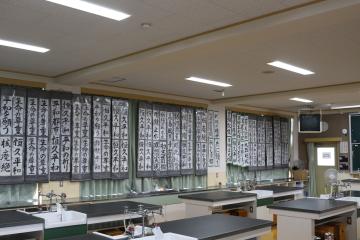 実験台と流しが並ぶ理科室の窓辺に大きな書道作品がずらりと掲示されている風景