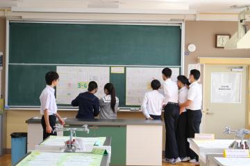 教室で数人の学生が黒板の前に集まり掲示物を見ている
