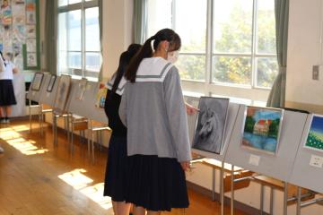 セーラー服の制服を着た女子生徒が教室の窓際で飾られた絵を眺めている