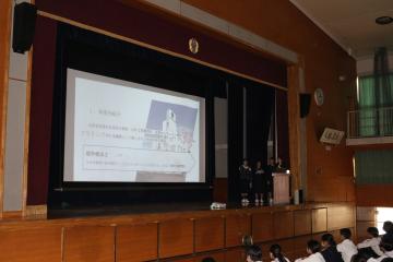 学校の講堂で壇上のスピーカーがスクリーンにスライドを投影し聴衆が席に座っている様子