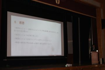 講堂の舞台で大きな投影スクリーンにスライドが表示され演台に立つ発表者がいる風景