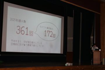 講堂でスクリーンに大きな数字361と172が表示されたスライドを映して右側の演台で発表者が立っている場面