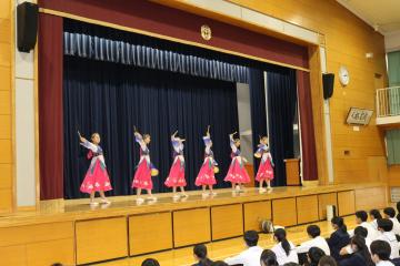 学校の講堂の舞台でピンクの衣装を着た数人の子どもたちが並んで踊りを披露している様子を客席の観客が見ている