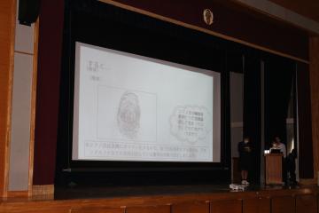 暗い講堂のステージでスクリーンにスライドが投影され右側の壇上で講演者が説明している様子