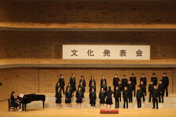 舞台上でピアノ伴奏に合わせて整列して合唱する学生たちが文化発表会の横断幕の前に並んでいる