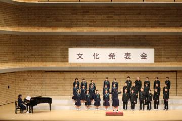 大きなホールの舞台でピアノ伴奏を行う演奏者と並んで立つ合唱団が横長の看板に文化発表会と書かれた背景の前で歌っている