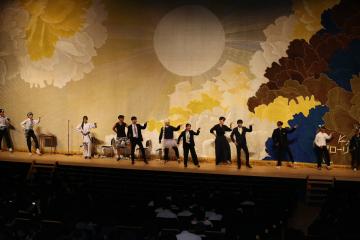 ステージ上で背景画の前に並んで歌い踊るバンドと数人のパフォーマーと楽器を演奏する人々