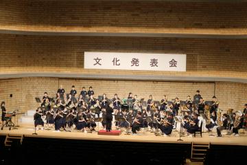 大勢の演奏者がステージに並び指揮者を中心に吹奏楽団が文化発表会の横断幕の下で演奏している光景