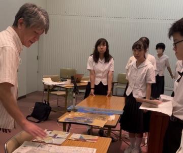 昨年度のワークショップの様子（教室のテーブルを囲んで大人と制服の学生たちが地図や資料を広げて説明を聞いたり確認したりしている場面）